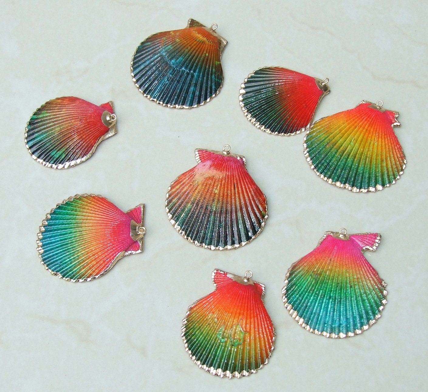 May include: Seven colorful seashells with gold trim. The seashells are painted with shades of red, orange, yellow, green, blue, and purple. The seashells are arranged in a circle on a white surface.