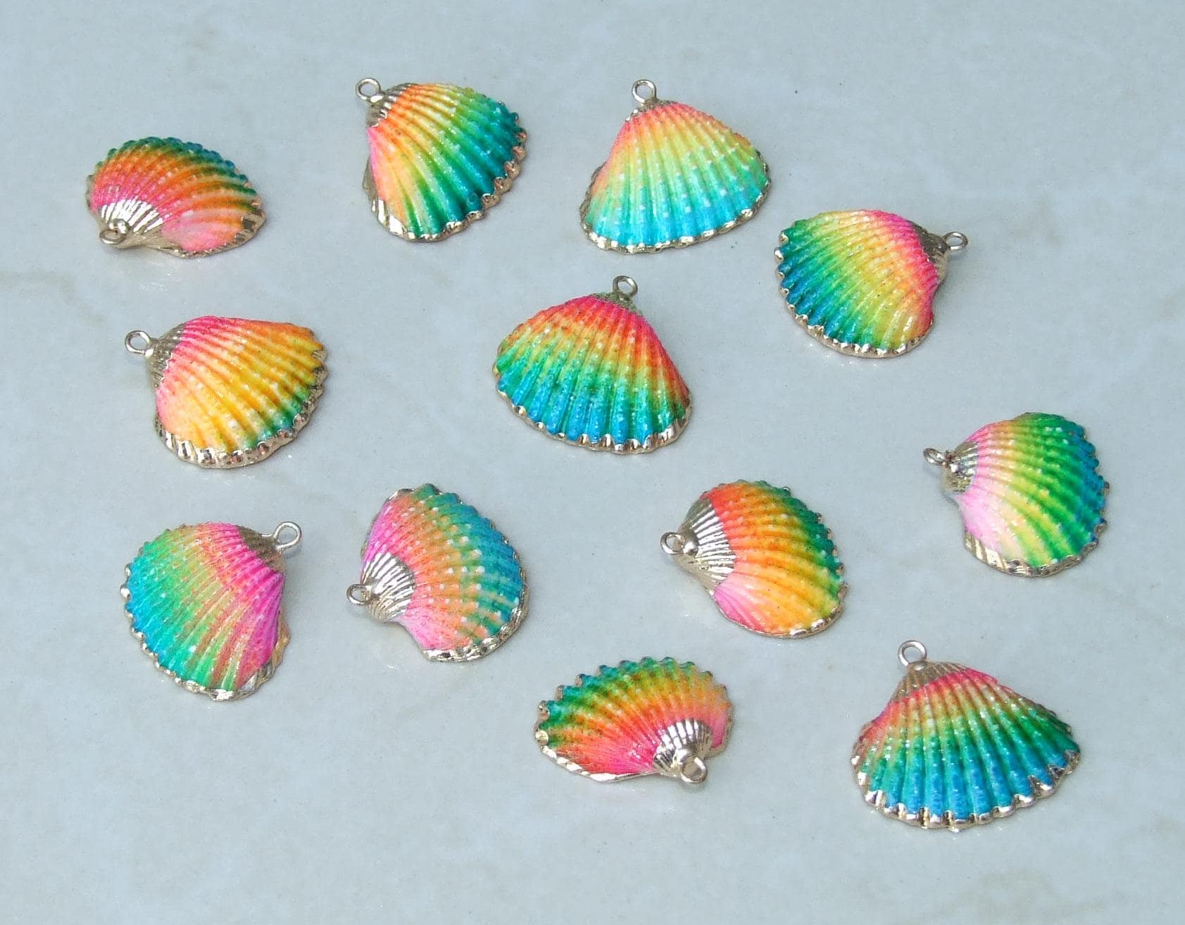 May include: A collection of twelve small seashell charms, each featuring a vibrant gradient of pink, yellow, and blue colors. Each charm has a textured, ribbed surface and a small metal loop at the top for attachment, arranged in a scattered pattern.